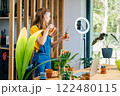 Beautiful woman blogger uses a ring lamp and phone, records video on how to care for succulents and how to prepare soil for them to replant at home in modern cozy environment. 122480115