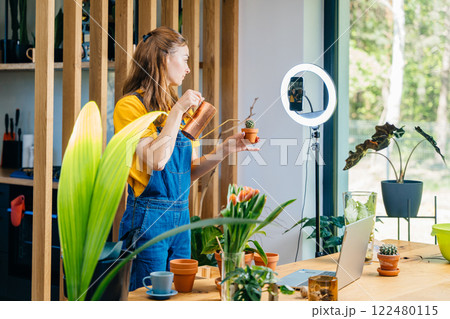 Beautiful woman blogger uses a ring lamp and phone, records video on how to care for succulents and how to prepare soil for them to replant at home in modern cozy environment. Beautiful woman blogger uses a ring lamp and phone, records video on how to care for succulents and how to prepare soil for them to replant at home in modern cozy environment. 122480115