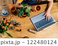 Top view close up of hands typing on notebook keyboard, wooden table with clay pots, plants, bouquet of tulips. 122480124
