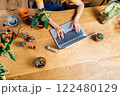 Top view close up of female hands on notebook keyboard on large wooden table with clay pots, plants, cactuses, succulents, bouquet of tulips. Woman botanist work from home. 122480129