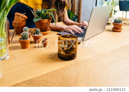 Unrecognizable young woman botanist hobbyist shopping for pots and soil for her hobby in online store at home. Close up of hands on notebook keyboard, wooden table with clay pots, plants,. 122480131