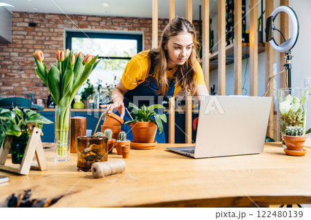 Young female botanist checking online orders on laptop. European woman entrepreneur working on her laptop in the living room on wooden table at cozy home interior. 122480139