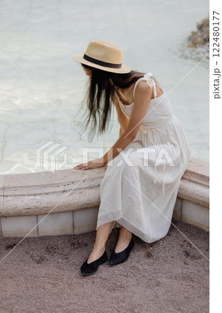 young Africa American woman in white dress straw hat sits white marble fountain and touches water young Africa American woman in white dress straw hat sits white marble fountain and touches water 122480177