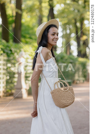 African American woman in white dress, hat walks in park poses against background of green hedge 122480178