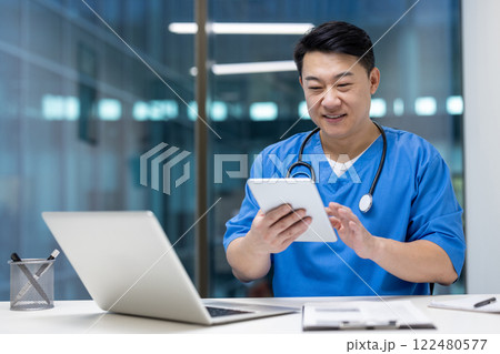 Asian man doctor using tablet and laptop in office, showcasing efficiency and modern healthcare solutions. Wearing scrubs, smiling, focused on digital screen, enhancing medical productivity. Asian man doctor using tablet and laptop in office, showcasing efficiency and modern healthcare solutions. Wearing scrubs, smiling, focused on digital screen, enhancing medical productivity. 122480577