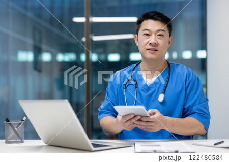 Asian man doctor in blue scrubs holds tablet in modern office, highlighting medical professionalism and technology. Stethoscope accents clinical expertise. Asian man doctor in blue scrubs holds tablet in modern office, highlighting medical professionalism and technology. Stethoscope accents clinical expertise. 122480584