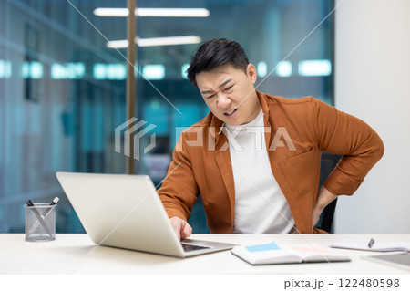 Asian businessman in office suffering back pain working on laptop. Man displays discomfort in office setting, highlighting strained posture and physical workplace stress. 122480598
