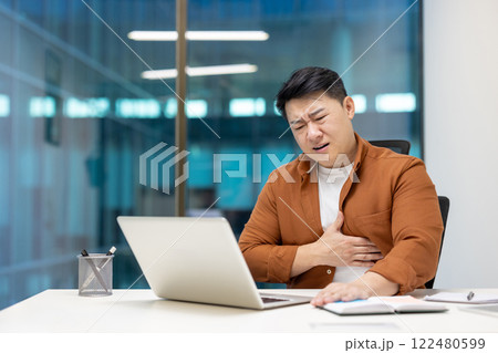 Mature Asian man feeling discomfort, placing hand on chest indicating possible heart attack at office desk with laptop. Concept of health, stress management, workplace. 122480599