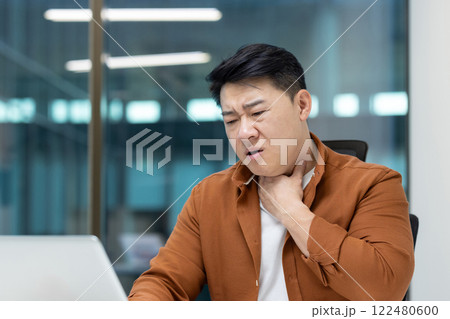Asian man experiencing pain in his throat while sitting in front of a laptop Asian man experiencing pain in his throat while sitting in front of a laptop 122480600