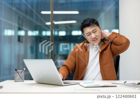 Asian man holding his neck with discomfort while seated at a desk with a laptop, illustrating workplace strain or health concerns related to prolonged sitting or poor posture in an office setup. 122480601