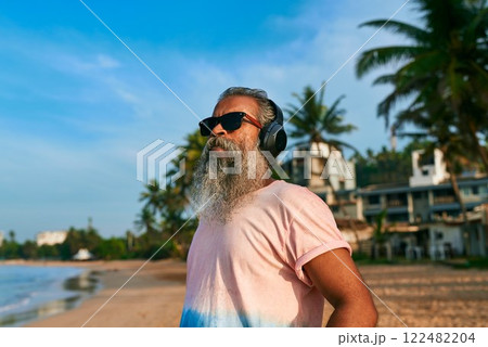 Grey-bearded senior man enjoying music on tropical beach. Stylish elder in sunglasses and wireless headphones. Relaxing, having fun by sea waves under palm trees. Elegant modern lifestyle. 122482204