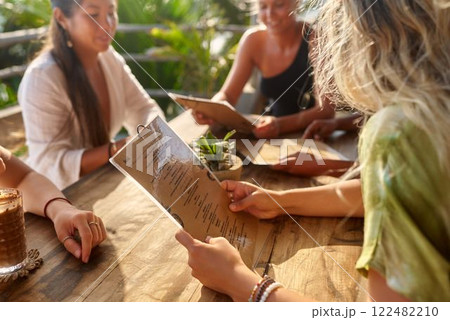 Diverse women meet at restaurant for brunch. Friends chat, choose food. Outdoor table, sunny day. Women club gathering, enjoying meal, drinks. Relaxed socializing, lifestyle scene. 122482210