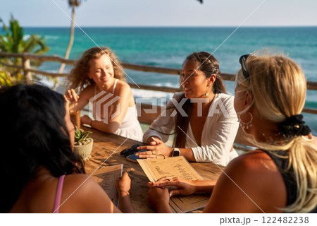 Diverse women in seaside restaurant enjoy sun. Friends choose dishes, drinks from menu. Relaxed group dining, discussing options, vacation, sunny weather, ocean view, casual atmosphere. 122482238