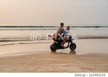 Couple rides motorbike on beach. Woman relaxes with legs up, man steers, enjoying leisure time by ocean. Romantic travel adventure in sunset light, beach vibes. Couple rides motorbike on beach. Woman relaxes with legs up, man steers, enjoying leisure time by ocean. Romantic travel adventure in sunset light, beach vibes. 122482239