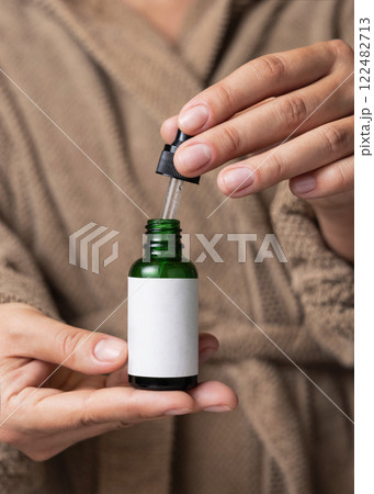 Hands in brown bathrobe opening green dropper bottle with label closeup, cosmetic mockup Hands in brown bathrobe opening green dropper bottle with label closeup, cosmetic mockup 122482713