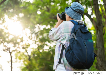 Portrait of Asian mature woman holding a camera and backpack behind her back, an Asia active senior woman enjoying nature in park. Standing on a trail in a forest outdoors. Enjoying active travel trip 122485696