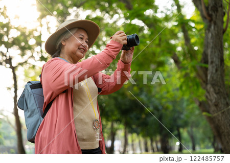 Portrait of Asian mature woman holding a camera and backpack behind her back, an Asia active senior woman enjoying nature in park. Standing on a trail in a forest outdoors. Enjoying active travel trip 122485757