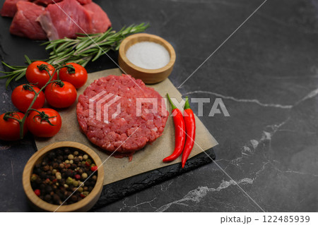 The formed minced meat cutlet lies on a paper backing on a dark marble background surrounded by spices, vegetables and rosemary, with chunks of beef meat in the background. The concept of cooking The formed minced meat cutlet lies on a paper backing on a dark marble background surrounded by spices, vegetables and rosemary, with chunks of beef meat in the background. The concept of cooking 122485939
