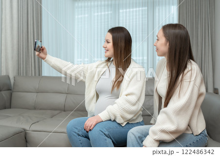 Young pregnant woman taking a selfie with her friend at home on a sofa, smiling, with copy space. Concept of friendship, maternity, capturing special memories during pregnancy. 122486342