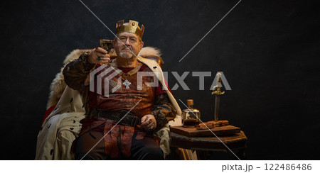Victory. Senior man, medieval king in crown sitting on throne and raisin glass of wine against black studio background 122486486