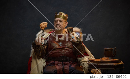 Senior man, medieval king in period attire sitting on throne with meat on sword and glass of wine against black studio background Senior man, medieval king in period attire sitting on throne with meat on sword and glass of wine against black studio background 122486492