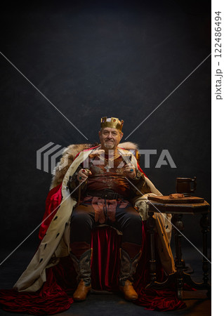 Senior man, medieval king in period attire sitting on throne with meat on sword and glass of wine against black studio background 122486494