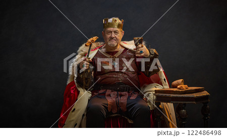 Senior man, medieval king in period attire sitting on throne with meat on sword and glass of wine against black studio background. Balance between power and pleasure. 122486498