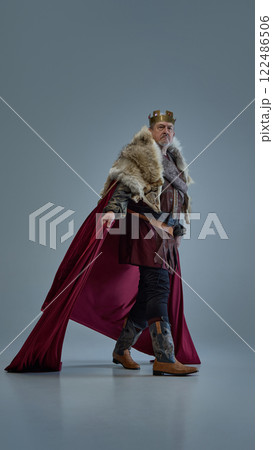 Elderly focused man, medieval king walks forward with determination, cloak flowing behind against grey studio background. 122486506