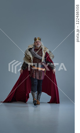 Man, medieval king walks forward with determination, cloak flowing behind against grey studio background. 122486508