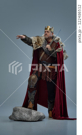 Full-length portrait of senior man as medieval king, in crown with sword, standing on stone and pointing forward against grey studio background 122486512