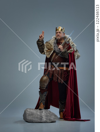 Senior serious man as medieval king standing on stone with sword and gesturing with one hand raised against grey studio background. Hope Senior serious man as medieval king standing on stone with sword and gesturing with one hand raised against grey studio background. Hope 122486513
