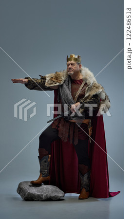 Commanding medieval king on rock, pointing forward with authority against grey studio background. Commanding medieval king on rock, pointing forward with authority against grey studio background. 122486518