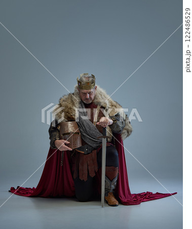 Serious man, medieval king with crown and fur cloak holding metal helmet and sitting on one knee with respect and honor against grey studio background Serious man, medieval king with crown and fur cloak holding metal helmet and sitting on one knee with respect and honor against grey studio background 122486529