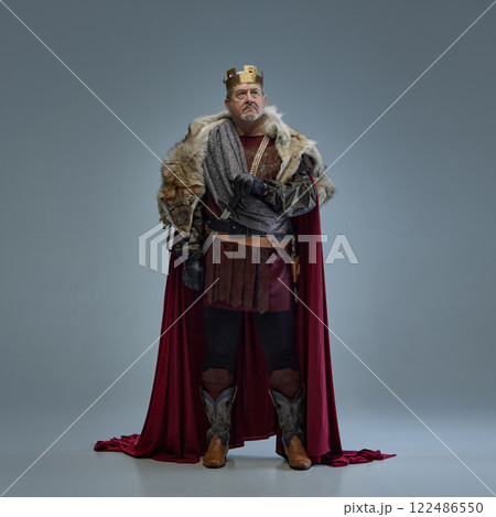 Full-length portrait of elderly bearded man, medieval king in fur-lined cloak and golden crown, standing with hands on swords. Authority and power Full-length portrait of elderly bearded man, medieval king in fur-lined cloak and golden crown, standing with hands on swords. Authority and power 122486550