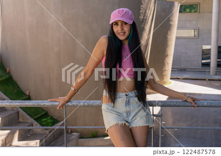 Young millennial smiling latina woman with a pink cap and long hair leaning on a railing 122487578