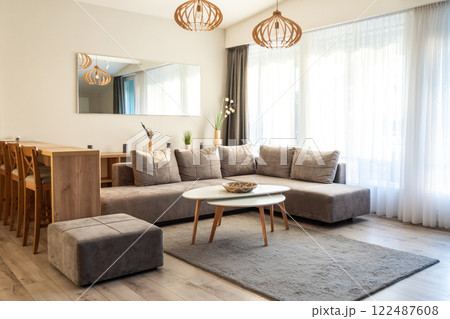 Bright living room with gray sectional sofa, white coffee table, wooden dining chairs, and large windows 122487608