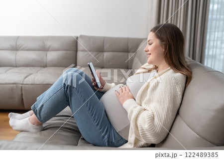 Pregnant woman reclining on a couch using smartphone for browsing and shopping, enjoying a moment of digital convenience. Concept of technology, e-commerce, maternity, and relaxation at home. Pregnant woman reclining on a couch using smartphone for browsing and shopping, enjoying a moment of digital convenience. Concept of technology, e-commerce, maternity, and relaxation at home. 122489385