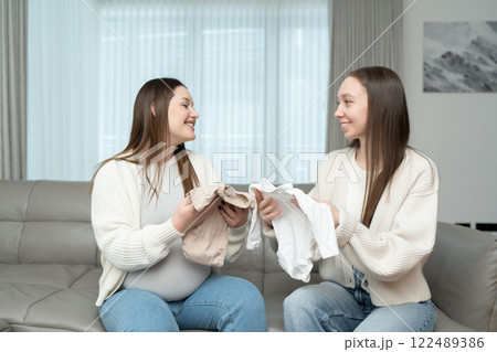 Young pregnant woman and her female friend looking at baby clothes while sitting on a couch. Concept of motherhood, friendship, preparation, excitement, and bonding. 122489386
