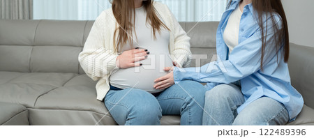 Banner of pregnant woman in third trimester sitting on sofa while her female friend touches her belly with care. Concept of maternity, friendship, connection, support and emotional bonding. 122489396