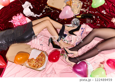 Women's legs tangled in Christmas lights rest on pink satin covered in spilled champagne, crushed confetti, and broken glasses, revealing reckless afterparty mood. 122489459
