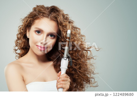 Friendly woman with curly hairstyle using hot curling tongs iron, indoor fashion portrait.  122489680