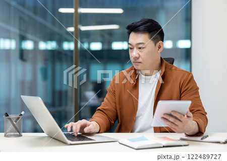 Serious focused asian man working with laptop inside office, mature man holding tablet computer in hands, typing on keyboard. Businessman inside office at workplace. Serious focused asian man working with laptop inside office, mature man holding tablet computer in hands, typing on keyboard. Businessman inside office at workplace. 122489727