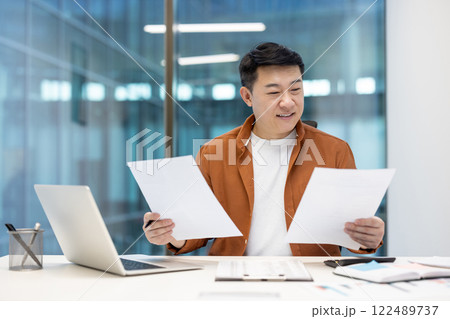 Successful Asian financier checking investment reports. Happy satisfied man smiling holding documents and papers in hands. Businessman at workplace inside office doing paperwork. 122489737