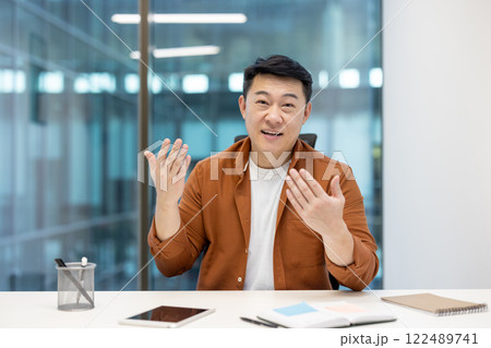Webcam view, video call, Asian man looking at camera, smiling and communicating with colleagues and partners remotely. Successful businessman working inside office at workplace. Webcam view, video call, Asian man looking at camera, smiling and communicating with colleagues and partners remotely. Successful businessman working inside office at workplace. 122489741