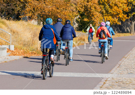 Group senior bikers pedal on paved path during vibrant autumn day, passing golden foliage and scenic Elbe riverside. People enjoy nature healthy air freedom while cycling together under sunny sky 122489894