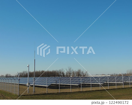 solar panels in agricultural field behind lattice fence with surveillance camera on sunny day, solar energy receiving equipment at station, photovoltaic panels in suburban area solar panels in agricultural field behind lattice fence with surveillance camera on sunny day, solar energy receiving equipment at station, photovoltaic panels in suburban area 122490172