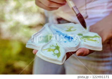 A close-up photo of a palette with watercolor paint and a brush in a woman's hand, drawing in nature in the forest. Horizontal background from the plein-air. 122491171