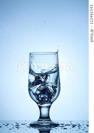 Refreshing splash of ice in a crystal-clear glass under serene blue backdrop Refreshing splash of ice in a crystal-clear glass under serene blue backdrop 122491543