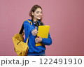 Front view of schoolgirl standing, looking at camera, smiling. Beautiful young teenage female with earphones and rucksack, showing super. Concept of education and youth. 122491910