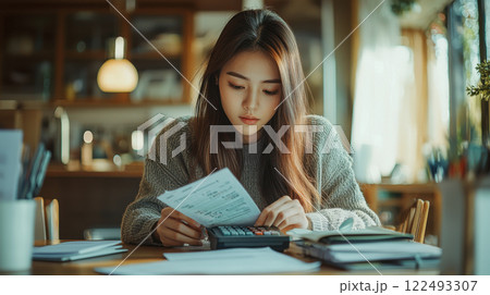 Young asian woman calculating expenses at home using a calculator in a cozy sunlit room with books and papers on wooden table 122493307